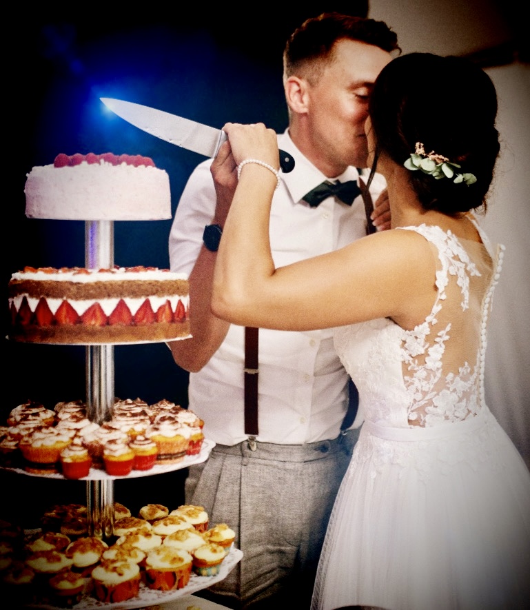 Romantischer Moment: Ein Brautpaar schneidet gemeinsam die mehrstöckige Hochzeitstorte an, die mit Cupcakes und frischen Früchten liebevoll dekoriert ist.