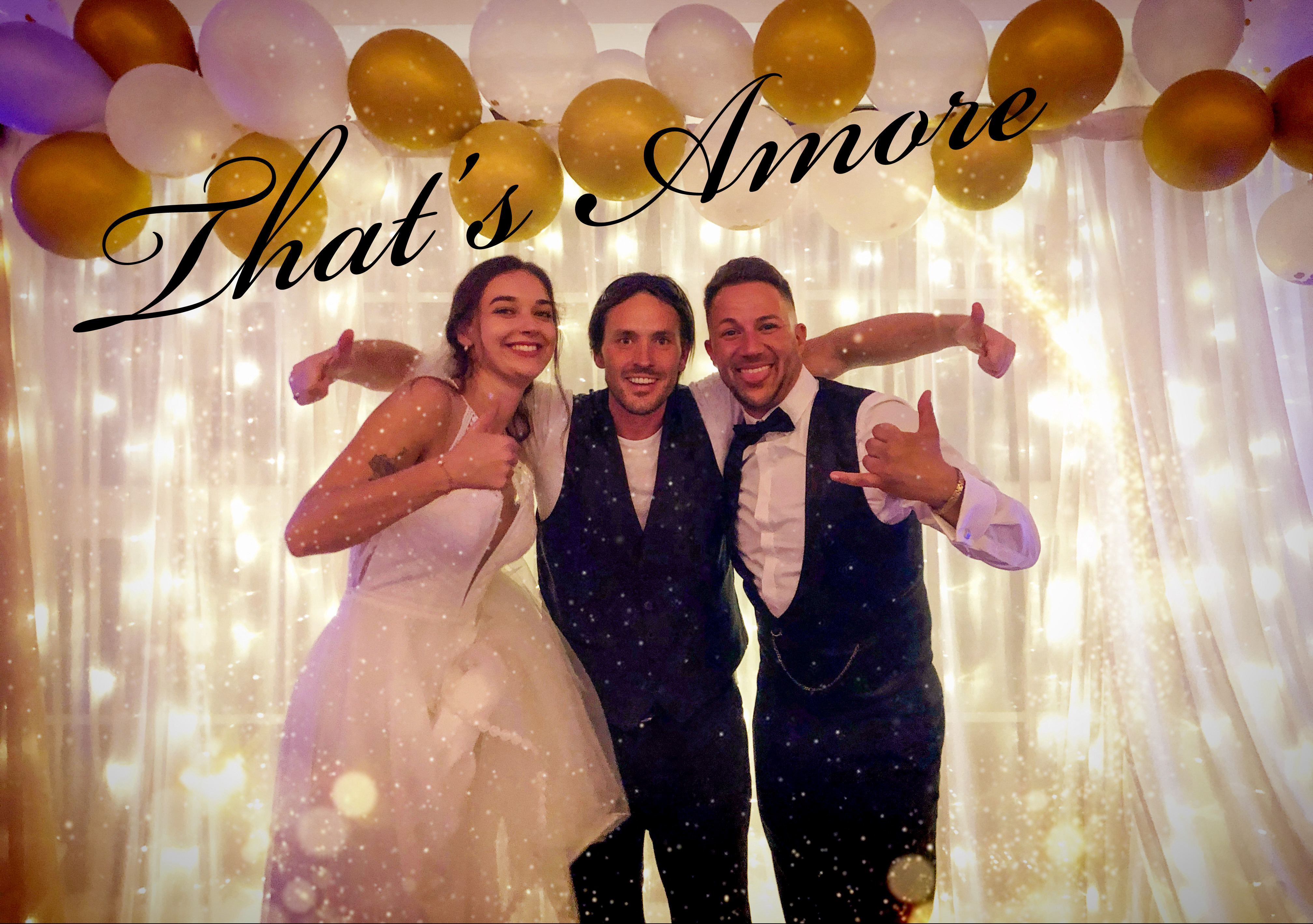 Ein glückliches Brautpaar posiert Arm in Arm mit ihrem Hochzeits-DJ Rico Klemm vor einer festlichen Lichterwand mit goldenen Ballons und dem Schriftzug That's Amore.